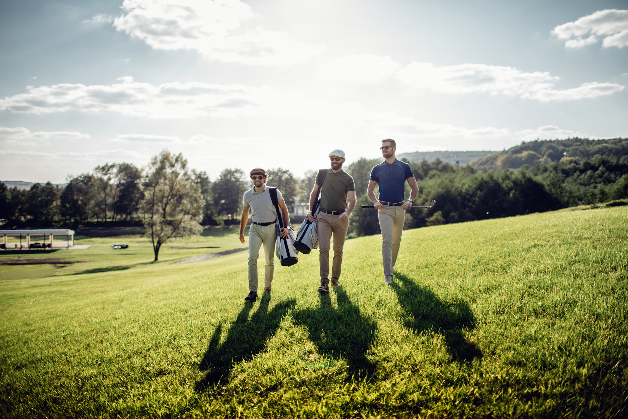 Three men walking across a golf course in the sunshine carrying their equipment